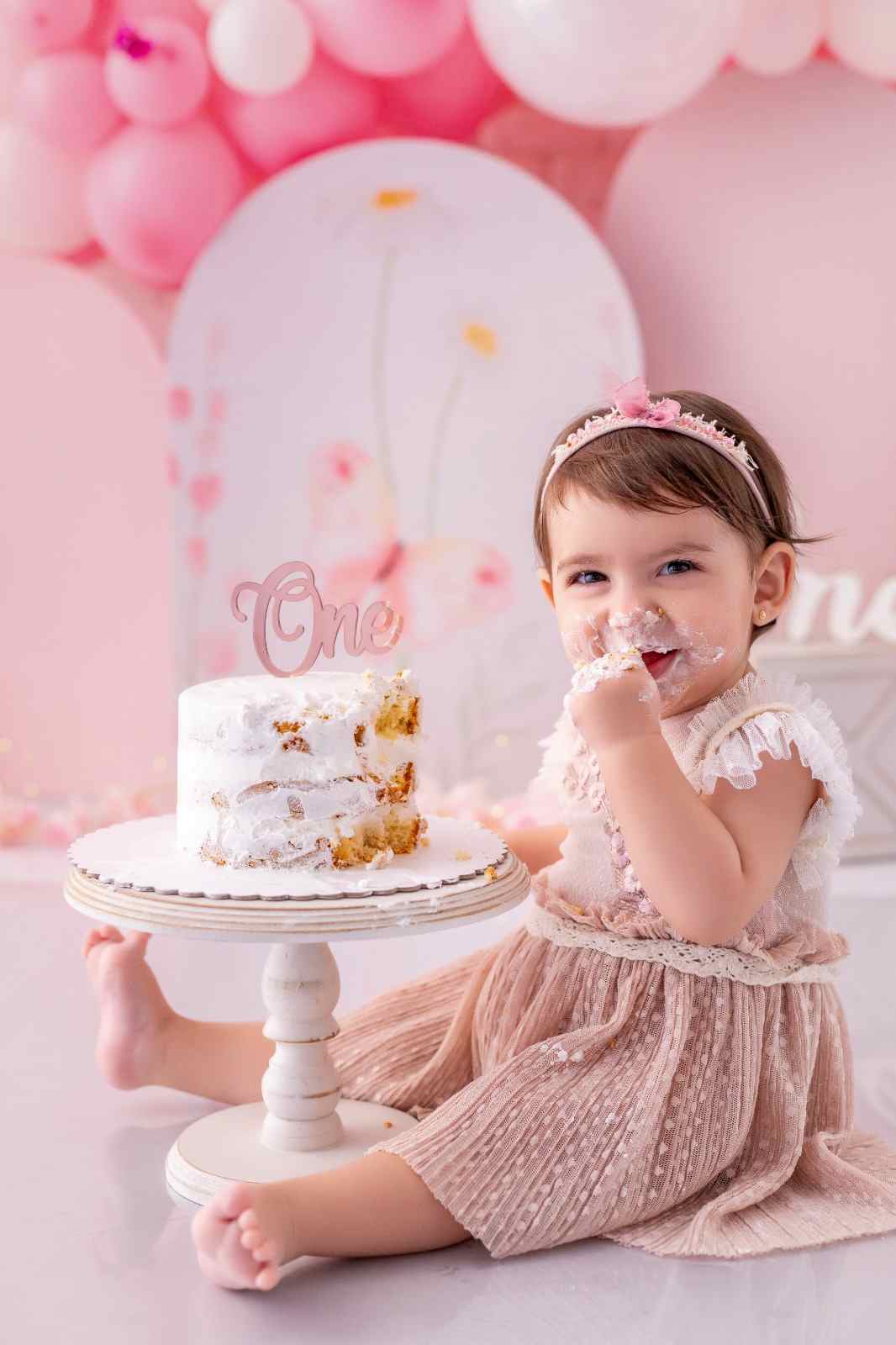 Bebê com vestidinho, em cenário todo rosa, comendo um bolo inteiro com as próprias mãos.