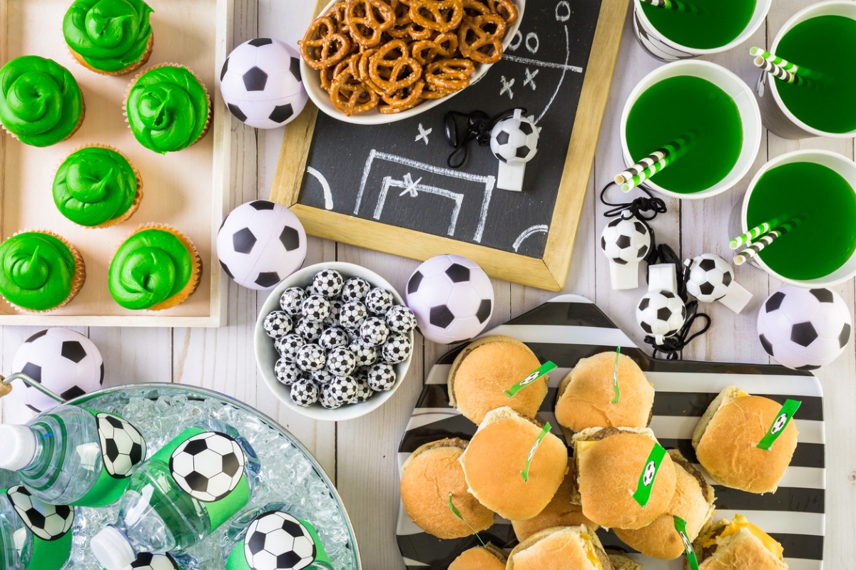 mesa decorada com tema de futebol para uma festa. A mesa inclui cupcakes com cobertura verde, salgadinhos, pretzels, copos com bebidas verdes, e várias bolas de futebol decorativas