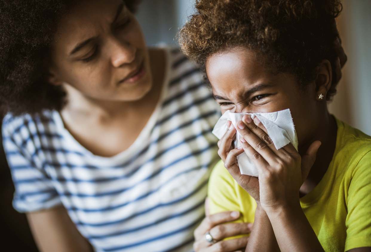 Criança assoando o nariz com um lenço enquanto um adulto, ao lado, observa com preocupação.