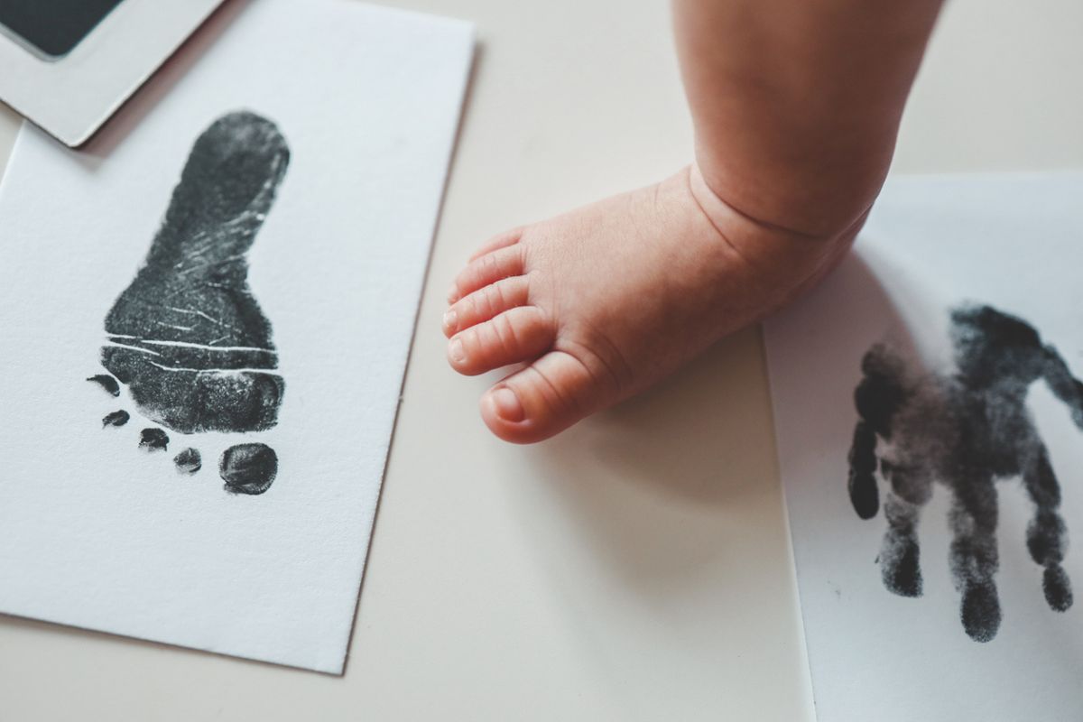 Pé de um bebê ao lado de impressões de tinta preta do pé e da mão do bebê em folhas de papel branco. 