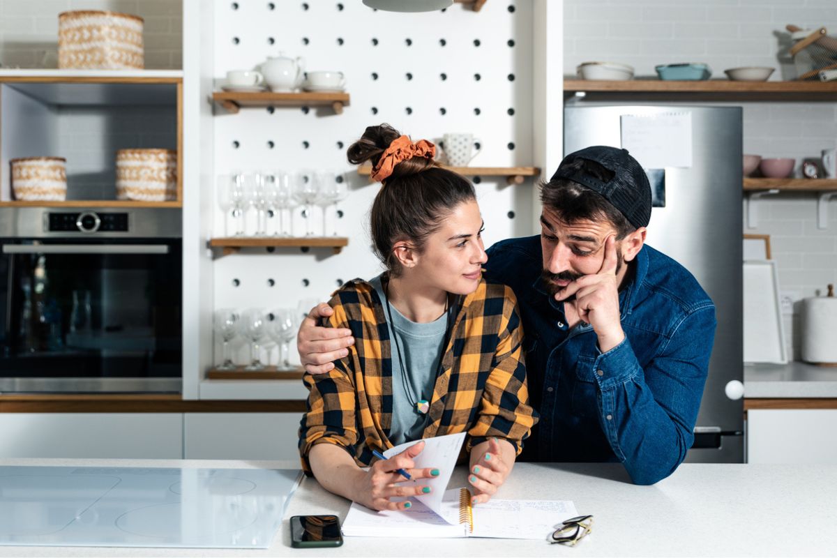 casal na cozinha, parecendo discutir finanças ou planejamento. A mulher está segurando um papel e uma caneta, enquanto o homem, com a mão no rosto, olha para o papel com uma expressão pensativa.