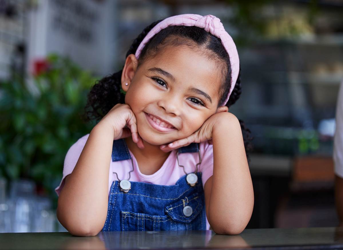 Menina sorridente com as mãos apoiadas no queixo e cotovelos na mesa, com acessório rosa no cabelo